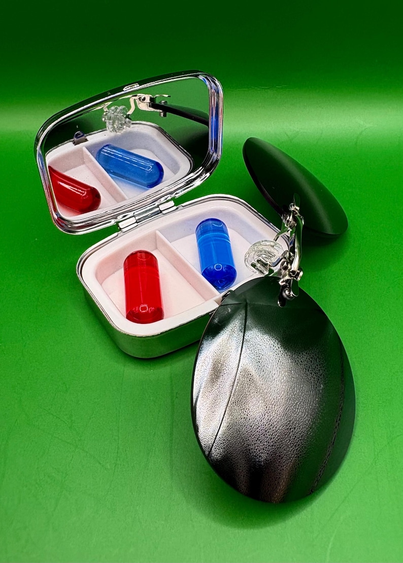 May include: A silver pill case with a mirror, containing red and blue capsules. A dark green, oval-shaped accessory hangs from the case. The background is a solid green.