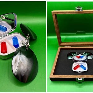 May include: A silver pill case with a mirror, containing a red and a blue capsule. The case is attached to a dark green, oval-shaped keychain. A wooden display box holds the case and capsules, with a black interior.