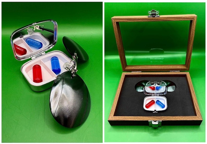 May include: A silver pill case with a mirror, containing a red and a blue capsule. The case is attached to a dark green, oval-shaped keychain. A wooden display box holds the case and capsules, with a black interior.