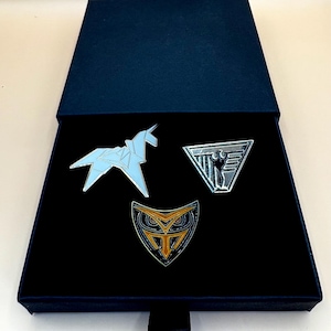 May include: A navy blue presentation box with three enamel pins. The pins include a light blue origami unicorn, a silver triangle with a figure, and an owl design with gold and black accents. The box has a black interior.