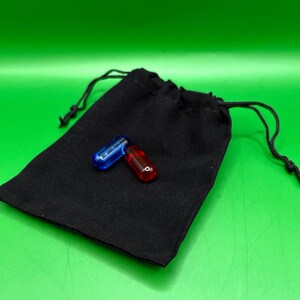 May include: A black drawstring bag with two capsule-shaped objects. One capsule is transparent blue, and the other is transparent red. The bag is made of a soft, dark fabric. The background is a solid green.