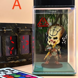 May include: A Predator figurine inside a clear display case with a black top. The figurine is yellow, brown, and black. A remote control with color options is in the foreground. A decorative panel is on the left.