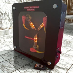 Puede incluir: Un accesorio negro y rojo con una ventana transparente que muestra tubos y cables brillantes. El texto "DISCONNECT CAPACITOR DRIVE BEFORE OPENING" y "SHIELD EYES FROM LIGHT" es visible. Un mando a distancia con llavero está delante.