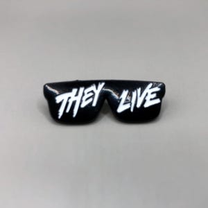 Könnte beinhalten: Schwarze Sonnenbrille mit weißem Text "THEY LIVE" auf den Gläsern.