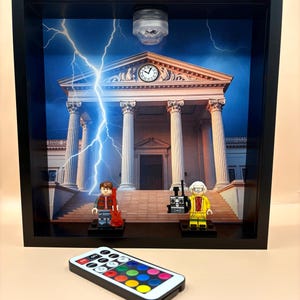 Può includere: Una scatola ombra incorniciata con personaggi Lego e uno sfondo ispirato a una scena cinematografica. La scena comprende un edificio con colonne, un orologio e fulmini. Un telecomando con opzioni di colore è in primo piano.