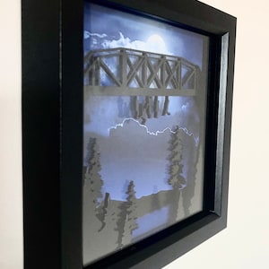 Puede incluir: Una caja de sombra enmarcada en negro con un dise&ntilde;o de papel recortado en 3D de un puente con personas caminando sobre &eacute;l. El fondo es un cielo nocturno azul y blanco con luna llena.