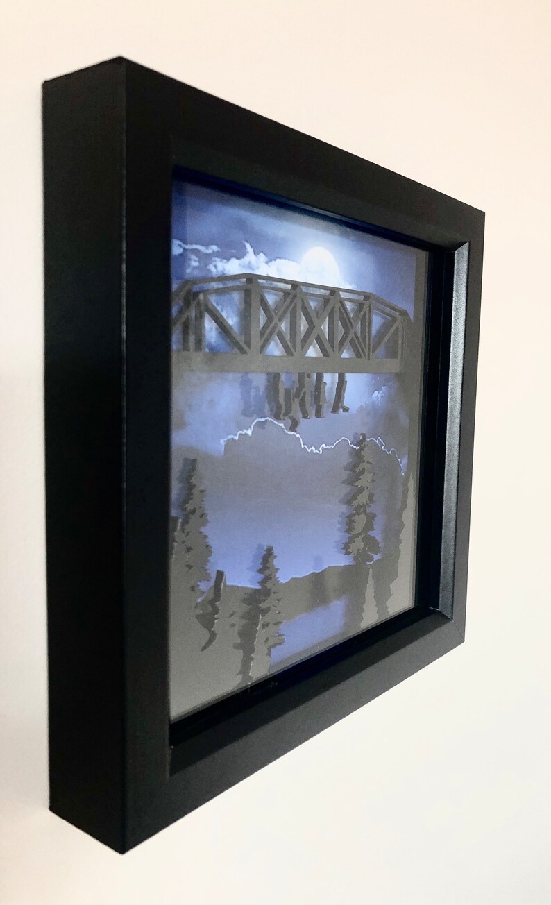 Puede incluir: Una caja de sombra enmarcada en negro con un dise&ntilde;o de papel recortado en 3D de un puente con personas caminando sobre &eacute;l. El fondo es un cielo nocturno azul y blanco con luna llena.