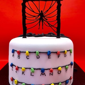 May include: A white cake with black decorations and colourful lights. The cake has the name "Sophia" on top, with a silhouette of a figure. The cake is decorated with colourful lights and letters.