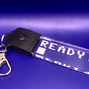 May include: A clear acrylic keychain with a black light-up button and the word "READY" in pixelated white letters.