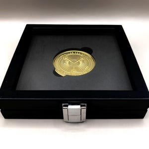May include: A gold-coloured coin featuring a design of two keys and a sunburst pattern is presented in a black velvet display case with a silver clasp.