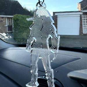 May include: Clear acrylic car air freshener in the shape of a figure, with etched contour lines. The charm is suspended by a black cord, hanging against a blurred background of a car interior and a residential area.