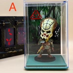 Peut inclure: Figurine Predator dans un présentoir transparent. La figurine porte un masque détaillé, des dreadlocks et tient un bâton. Le présentoir repose sur une base verte, avec une toile de fond de jungle et un symbole triangulaire rouge. La lettre "A" est dans le coin supérieur gauche.