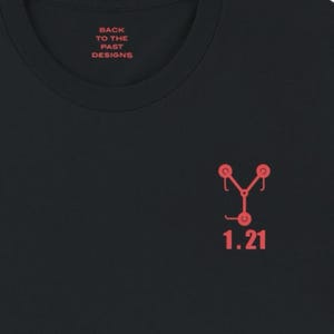 Könnte beinhalten: Schwarzes T-Shirt mit einer roten Grafik des "Fluxkompensators" aus dem Film Zurück in die Zukunft. Der Text "1.21" ist unter der Grafik gedruckt. Der Text "Back to the Past Designs" ist auf dem Nackenlabel gedruckt.