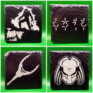 May include: Set of four black slate coasters with white graphic designs. Designs include a silhouette of a creature, symbols, a weapon, and a helmet. Each coaster is square and has a rough, textured surface.