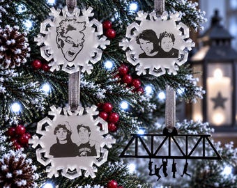 May include: Three clear acrylic snowflake ornaments featuring black etched portraits of characters and a black acrylic ornament depicting a bridge structure with silhouettes of four figures hanging from it, each suspended by a grey ribbon.