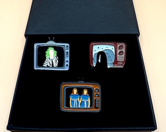 Puede incluir: Una caja negra que muestra tres pines de esmalte con diseños de televisión. Los pines presentan personajes de una película. Un pin muestra un personaje con cabello verde, otro muestra un personaje en un entorno oscuro y el último muestra dos personajes con vestidos azules.