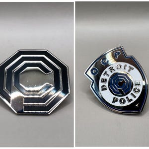 Puede incluir: Dos pines metálicos. Uno tiene forma de octágono con centro plateado y borde negro, con una gran letra "C". El otro tiene forma de escudo con el texto "DETROIT POLICE" y un diseño azul y blanco.