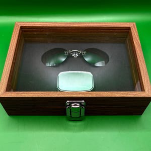May include: A wooden display case with a hinged lid, showcasing a pair of oval sunglasses and a small, rectangular, silver-colored case. The sunglasses have a silver bridge and dark lenses. The case is lined with black fabric.