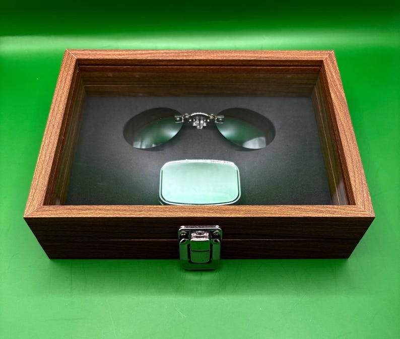 May include: A wooden display case with a hinged lid, showcasing a pair of oval sunglasses and a small, rectangular, silver-colored case. The sunglasses have a silver bridge and dark lenses. The case is lined with black fabric.