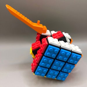 May include: A multi-coloured 3x3 Rubik's Cube made of plastic building blocks. The cube is in a partially solved state, with some blue, red, white, and black blocks visible. The cube has a bright orange lever attached to the top.