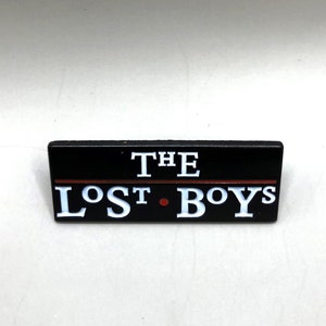 May include: Black enamel pin with white lettering that reads "THE LOST BOYS" with a red line above the text.