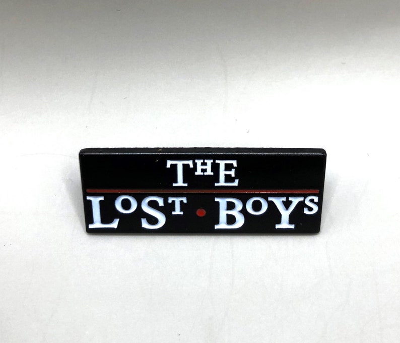 May include: Black enamel pin with white lettering that reads "THE LOST BOYS" with a red line above the text.