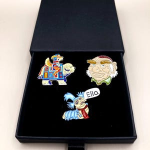Puede incluir: Tres pines de esmalte en una caja negra. Los pines representan personajes de la película Labyrinth. El primer pin es una criatura multicolor que monta un perro blanco. El segundo pin es un hombre con barba y una camisa verde. El tercer pin es una oruga azul con una bufanda roja y la palabra "Ello" en un globo de diálogo.