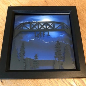 Puede incluir: Una caja de sombra enmarcada en negro con un dise&ntilde;o de papel recortado en 3D de un puente con figuras colgando de &eacute;l. El fondo es azul oscuro con una luna blanca y nubes. Las figuras est&aacute;n silueteados contra el fondo azul.