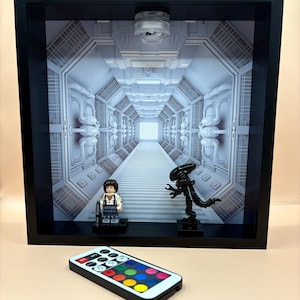 May include: A framed diorama featuring a futuristic corridor scene with a Lego figure and an Alien figure. The scene is lit with a bright light. A remote control with color options sits in front of the frame.
