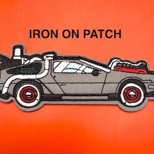 Op de afbeelding: Geborduurde opstrijkbare patch met een grijze DeLorean auto met witte en rode accenten. De patch heeft een zwarte rand en de tekst "IRON ON PATCH" in zwarte letters.