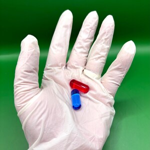 May include: Two capsules, one red and one blue, rest on a white gloved hand against a green background. The red capsule is oblong, while the blue one is cylindrical. The capsules are likely pharmaceutical products.