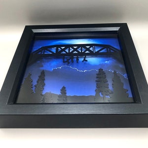 Puede incluir: Una caja de sombra enmarcada en negro con un fondo azul y una silueta negra de un puente con personas colgando de &eacute;l. El puente est&aacute; iluminado desde atr&aacute;s, creando un efecto de brillo. La escena se desarrolla sobre un fondo de &aacute;rboles y nubes.