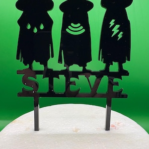 Puede incluir: Adorno para tarta de acrílico negro con tres figuras estilizadas con capas y sombreros, sobre el nombre "STEVE". Las figuras tienen diseños únicos, incluyendo gotas de lluvia, un símbolo wifi y rayos. El adorno está diseñado para insertarse en una tarta.