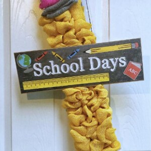 May include: A handmade pencil-shaped wreath with a sign that reads "School Days." The wreath is made of yellow, pink, gray, white, and black burlap. The sign has a globe, crayons, a pencil, a ruler, and an ABC book.