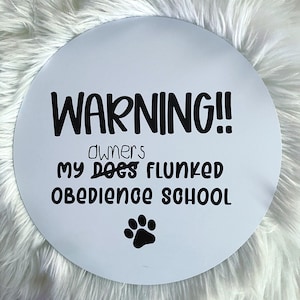 May include: A white, round sign with black text that reads "WARNING!! owners MY DOGS FLUNKED OBEDIENCE SCHOOL" and a black paw print. The sign is likely for a pet owner.