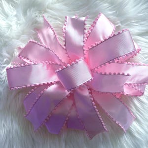 May include: A large, light pink bow made of satin ribbon. The edges of the ribbon are trimmed with small, matching pink pom-poms. The bow is arranged in a circular shape, with multiple loops and tails.
