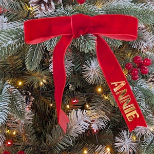 Op de afbeelding: Een helderrode fluwelen strik met de naam "ANNIE" in gouden letters, hangend aan een kerstboom. De strik is een feestelijke decoratie, die een vleugje elegantie toevoegt aan de kerstversiering. De boom is versierd met lichtjes en bessen.