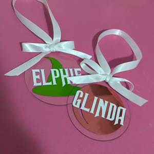 Op de afbeelding: Twee heldere, ronde ornamenten met witte tekst en witte strikken. Eén ornament heeft een groene heksenhoedafbeelding en de naam "ELPHIE". De andere heeft de naam "GLINDA" op een koperkleurige achtergrond. De ornamenten liggen op een roze ondergrond.