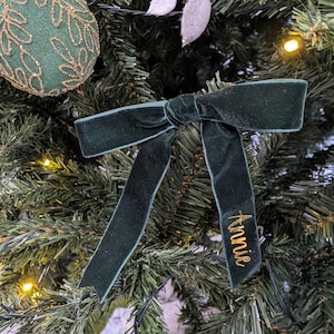 Op de afbeelding: Een donkergroene fluwelen strik met de naam "Annie" geborduurd in gouddraad, hangend aan een kerstboom. De strik is in het midden geknoopt en heeft lange uiteinden. De boom is versierd met ornamenten en lichtjes.