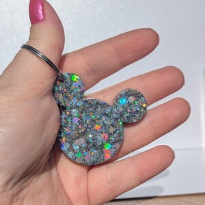 Op de afbeelding: Een zilveren glitter Mickey Mouse sleutelhanger met iriserende glitter. De sleutelhanger is bevestigd aan een zilveren ring.