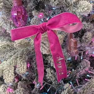 Op de afbeelding: Een helderroze fluwelen strik kerstboomversiering met de naam "Annie" geborduurd in zilveren letters. De strik is geknoopt en heeft lange, vloeiende uiteinden. De achtergrond toont een bevroren kerstboom.