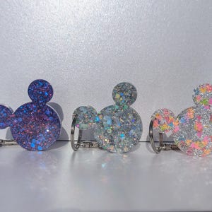 Op de afbeelding: Drie sleutelhangers in de vorm van Mickey Mouse-koppen. De sleutelhangers zijn gemaakt van helder hars gevuld met glitter. De eerste sleutelhanger is blauw met iriserende glitter, de tweede is zilver met iriserende glitter en de derde is roze met veelkleurige glitter.