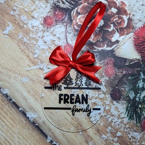 Op de afbeelding: Transparante, ronde kerstbal met de tekst "The FREAN family" in het zwart. De bal heeft een rood lint en strik om op te hangen. Het ontwerp bevat drie gestileerde kerstbomen.