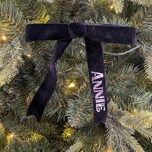 Op de afbeelding: Een zwarte fluwelen strik met lange linten, hangend aan een kerstboom. Het woord "ANNIE" is in zilver op een van de linten gedrukt. De strik is een feestelijke vakantieversiering.