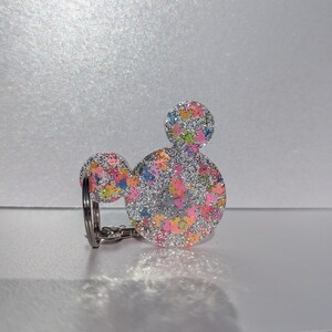 Op de afbeelding: Een zilveren glitter sleutelhanger in de vorm van Mickey Mouse oren met een kleurrijk confetti patroon.