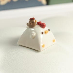 May include: A white ceramic chicken figurine with a small brown bird perched on its back. The chicken has red and yellow accents on its beak and comb.