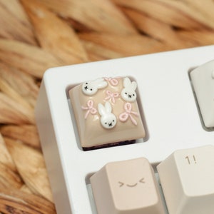 May include: A white mechanical keyboard with a custom keycap featuring a beige background with pink bows and white bunny illustrations. The keycap is labeled with the number "1!" and is surrounded by other keys with symbols like "< >" and "2@".