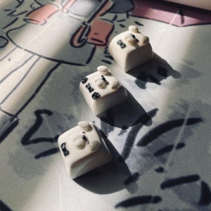 Puede incluir: Tres teclas blancas con detalles negros y un pequeño diseño negro en cada una. Las teclas están sobre una superficie de tela blanca con marcas negras.