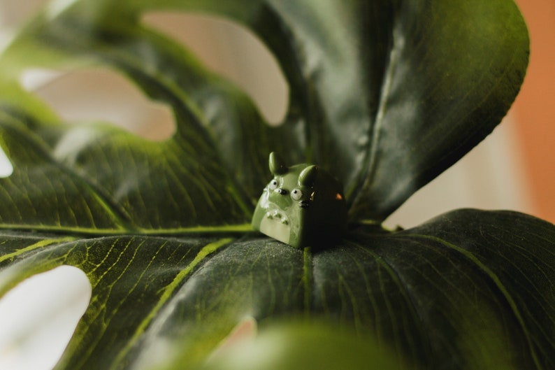 Puede incluir: Una figurita de cer&aacute;mica verde de un personaje de dibujos animados con ojos y orejas grandes, sentada sobre una hoja verde grande.