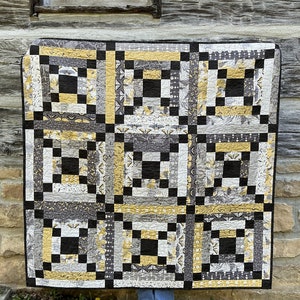 May include: A black, white, and gold quilt with a geometric pattern. The quilt is hanging on a wooden wall.